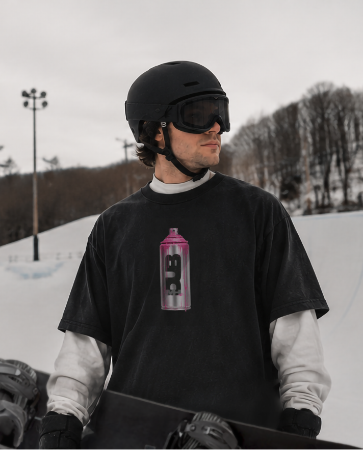 Person wearing MHDUB T-shirt, a black helmet and goggles, holding a snowboard on a snowy slope.